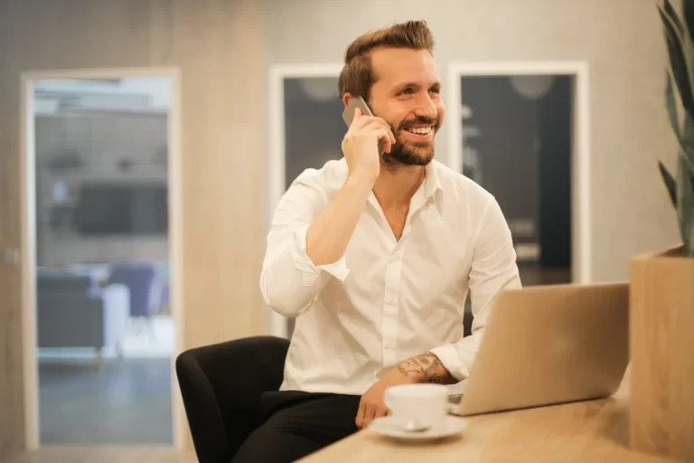 A smiling man with a beard and white shirt talks on a phone, sitting at a desk with a laptop and coffee.
