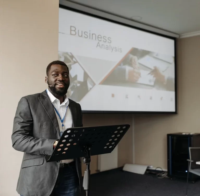 A smiling Black man in a suit stands behind a music stand with a "Business Analysis" presentation on a screen.